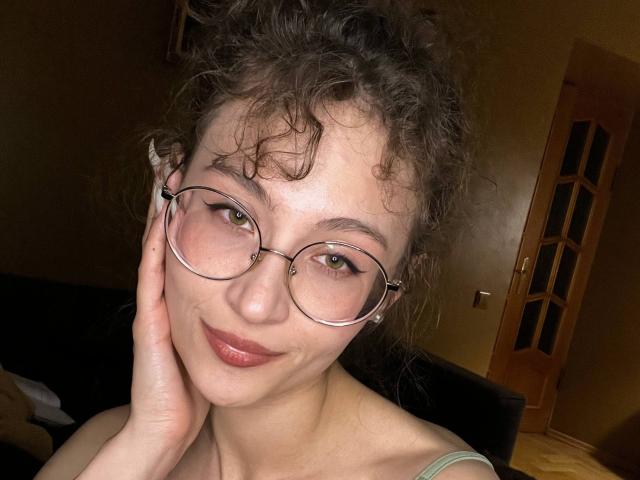 Alyce Erway, mit lockigem, braunem Haar, trägt eine Brille, hat helle Haut und ein leichtes Lächeln. Sie berührt ihre Wange mit einer Hand und zeigt dabei ihre grünen Augen und ihre rosafarbenen Lippen..