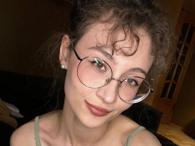 AlyceErway with curly brown hair, glasses, and pink lipstick, smiling slightly, wearing a green tank top, showing her face and upper chest. Her small tits are visible.