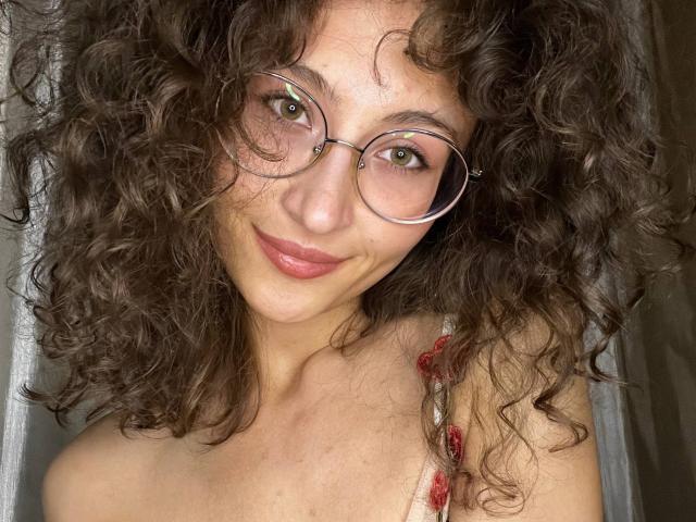 AlyceErway hat lockiges, braunes Haar, grüne Augen und rosafarbene Lippen. Sie trägt eine runde Brille und ein rotes Oberteil mit Trägern und lächelt sanft. Ihre Brüste sind teilweise sichtbar..
