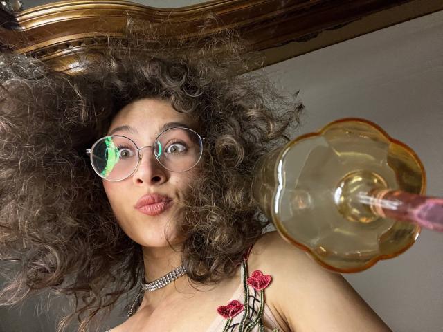 AlyceErway with curly brown hair, glasses, and a choker, pouting her lips, wearing a floral top with red roses, holding a yellow glass lamp with a pink handle.