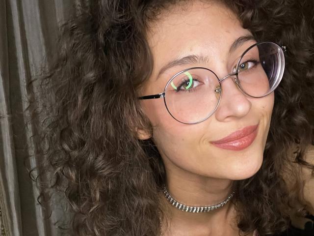 AlyceErway, met krullend bruin haar, draagt ronde brillen en een zilveren ketting. Ze heeft een klein neuspiercing en glimlacht met lichtroze lippen. Haar borsten zijn niet zichtbaar..