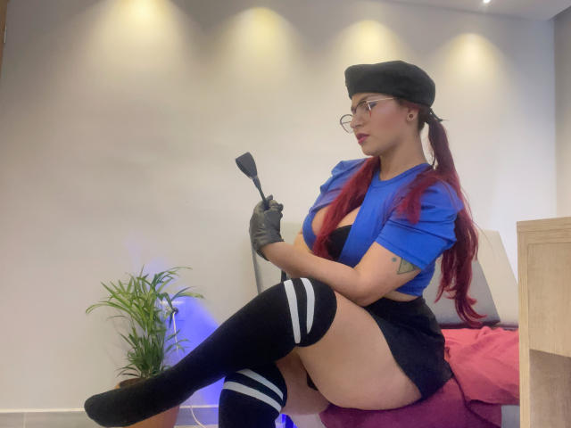 TrinitixPandora, wearing glasses, black beret, blue crop top, black shorts, thigh-high socks, holding a black whip, with red hair, sitting on bed, showcasing her large tits.