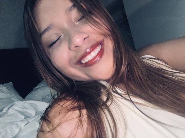 HeraJuno, sorridente con gli occhi chiusi, ha lunghi capelli castani, un piercing al naso e rossetto rosso. Indossa una maglietta bianca che lascia scoperta una spalla. I suoi denti sono bianchi e lucidi..