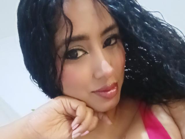MariaCatalina, met nat zwart krullend haar, roze lippenstift en donkere oogmake-up, leunt met haar kin op haar hand, draagt een roze top en laat haar kleine borsten zien..