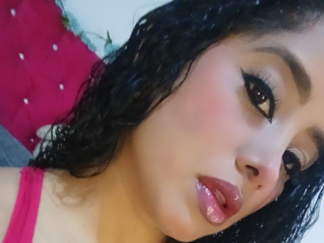 O rosto de MariaCatalina, com lábios rosados e brilhantes, cabelos cacheados escuros e delineador preto marcante. Ela está vestindo uma blusa rosa, olhando levemente para o lado com uma expressão suave..
