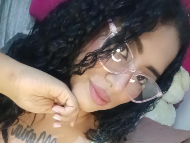 MariaCatalina with curly black hair, wearing pink glasses, pink lipstick, and a tattoo on her chest, touching her neck, showing her tits. Soft, cute, and sexy.