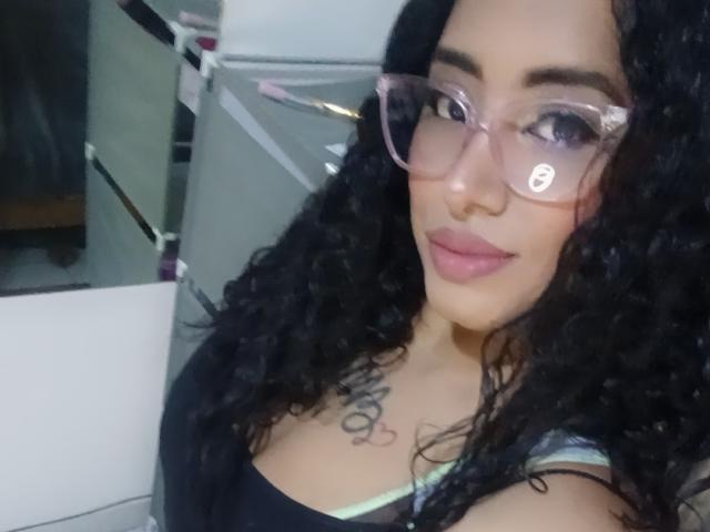 MariaCatalina, com cabelos pretos encaracolados, usando óculos rosa, tem uma tatuagem no peito e está mostrando os seios em uma selfie. Ela tem batom rosa e um leve sorriso..