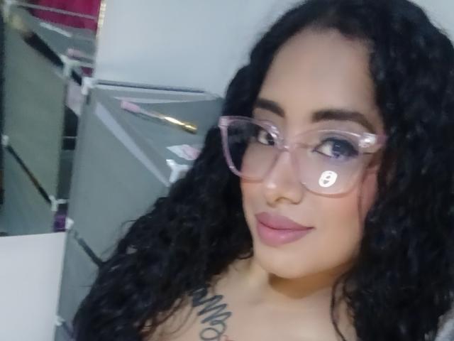 MariaCatalina with curly black hair, pink glasses, and pink lipstick, showing a tattoo on her chest, smiling with her tits slightly visible. She has a cute, sexy look.