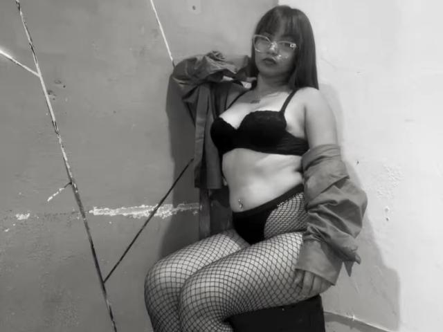EstefyChaud mit dunklem BH, schwarzen Unterhosen, Netzstrümpfen und offener Jacke, sitzend an einer rissigen Wand, mit Brille, die ihre Brüste und ihren Hintern zeigt, auf einem Schwarz-Weiß-Foto..
