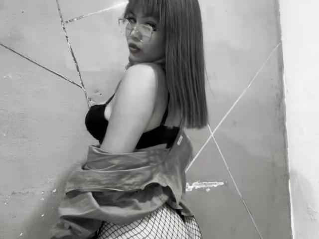 EstefyChaud con glúteos medianos, pantalones de rejilla, sujetador negro y chaqueta quitada, mirando por encima del hombro, usando gafas, en una foto en blanco y negro..