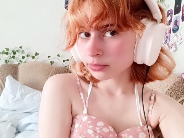 MireilleSpotwood, avec des cheveux roux, un haut floral rose, des écouteurs blancs, une peau pâle, de petites seins, des lèvres roses et douces, regardant la caméra, assise sur un canapé beige, avec un coussin bleu légèrement désordonné..