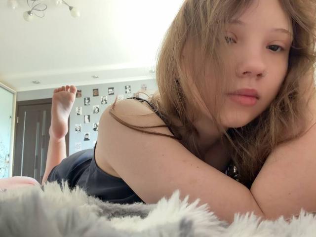 MarceneMalatesta, avec de longs cheveux bruns, un anneau de nez, allongée sur un lit moelleux, portant un haut noir, les jambes relevées, regardant la caméra, lumière douce, chambre à coucher, expression détendue, mignonne, sexy, ambiance bohème..