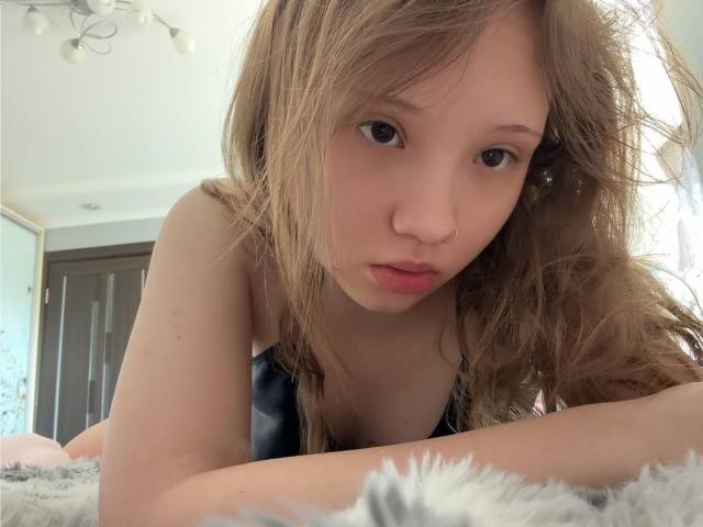 MarceneMalatesta a les cheveux bruns clairs et ondulés, et elle est penchée en avant avec les lèvres pincées, montrant de petites seins, et elle porte un piercing au nez, dans une chambre avec une couverture grise moelleuse..