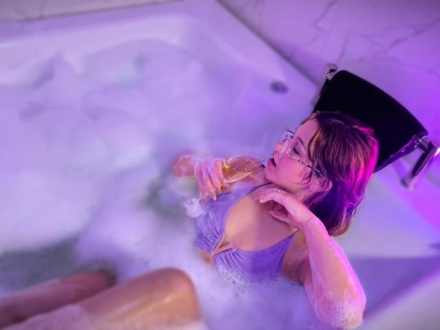 Elodie Gardner, con i capelli castani bagnati, gli occhiali e una canottiera viola, tiene una birra in una vasca bianca piena di bolle, illuminata da luci viola e rosa. Sembra rilassata, con un'aria sensuale..