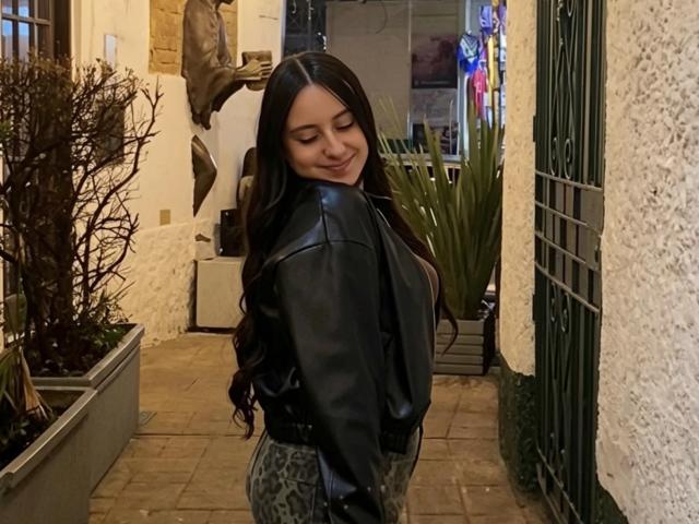 RosalieDupont with long black hair, wearing a black leather jacket and leopard print pants, smiling with closed eyes in a narrow, dimly lit, outdoor walkway.