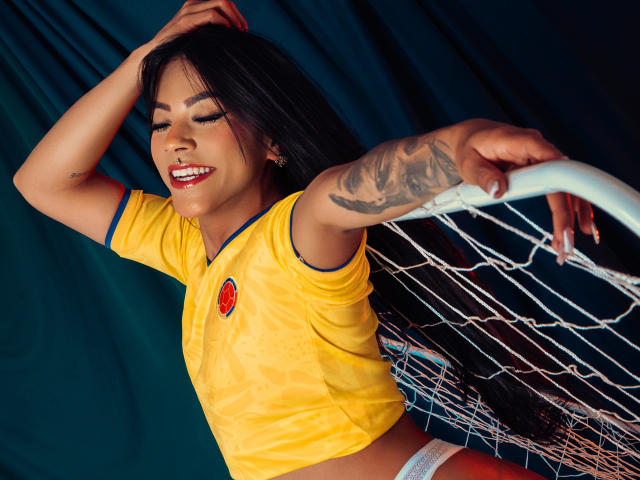 Amberv, avec ses longs cheveux noirs, souriante, portant un maillot de foot jaune, des sous-vêtements blancs, et tenant un filet de foot, a des tatouages sur son bras droit, et pose avec assurance..