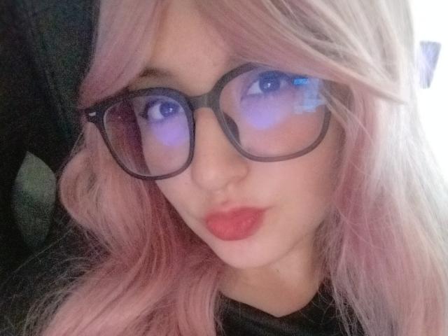 Susan Monroy, con i capelli rosa, gli occhiali neri e il rossetto rosso, guarda nella fotocamera con un'espressione leggermente corrucciata. I suoi occhi azzurri riflettono una leggera luce blu..