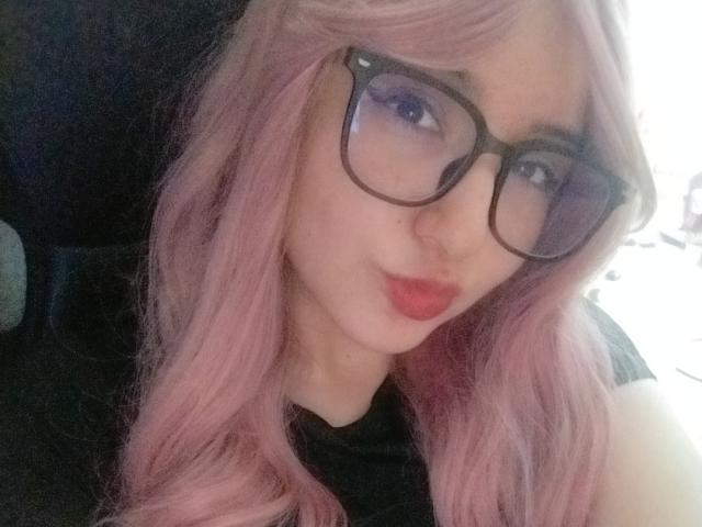 SusanMonroy, com cabelo rosa, óculos pretos e lábios vermelhos, mostrando um rosto fofo, vestindo uma blusa preta, com o traseiro não visível. Luz suave, sorriso discreto, selfie casual..