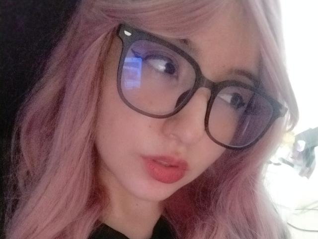 Susan Monroy, met roze haar, zwarte bril, volle roze lippen en een zwarte top, kijkt lichtelijk opzij, en haar reflectie is zichtbaar in het glas..