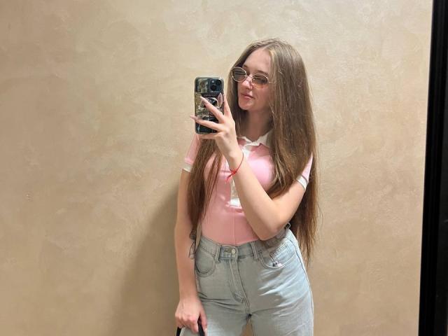 PamelaXu with long brown hair, glasses, pink shirt, white collar, light blue jeans, sticking tongue out, holding phone, mirror selfie, in a room with beige wall.
