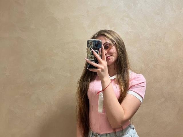 PamelaXu met lang bruin haar, een roze shirt met witte accenten, een bril, die een telefoon vasthoudt, een selfie maakt, lachend, een roze armband, een beige muur met textuur, een geruite rok, schattig, casual..