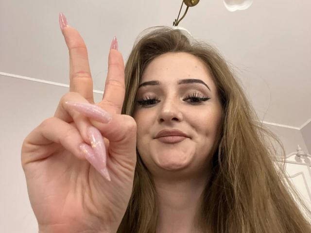 Pamela Xu mit langem, braunem Haar, rosafarbenen Nägeln und einem schmollenden Gesicht, die einen rosafarbenen, glitzernden Finger hochhebt und eine "pssst"-Geste macht, lächelnd mit Eyeliner..