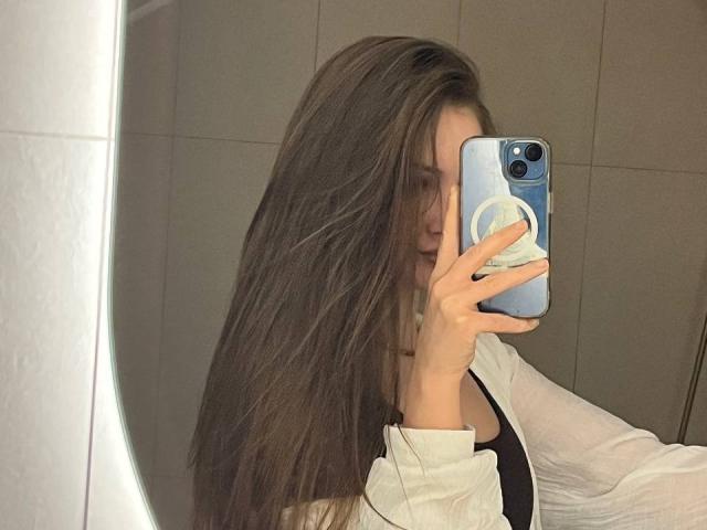 PamelaXu, com cabelos castanhos longos, tirando uma selfie no espelho, segurando um telefone azul com um suporte popsocket branco, vestindo uma camisa branca, mostrando seus seios..
