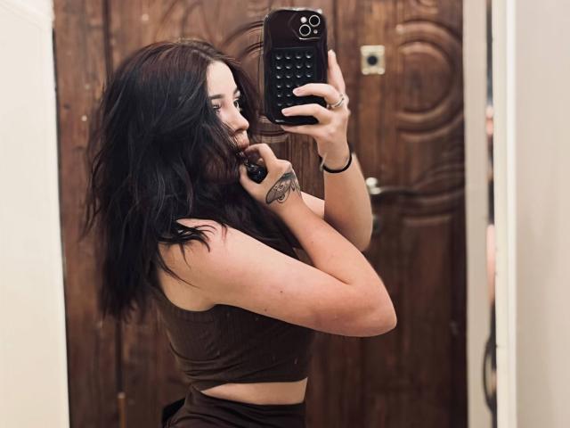 SinfulKitty with dark wavy hair, brown tank top, holding phone, showing tattoo on right wrist, taking mirror selfie, wood door in background, slightly biting phone case.