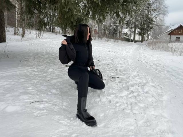 SinfulKitty ajoelha-se na neve, vestindo um casaco preto, calças e botas, com uma mochila preta e segurando uma sacola. Ela tem cabelo escuro e uma figura esguia e atraente..