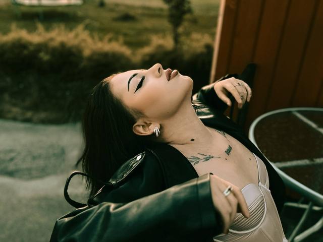 SinfulKitty con i capelli scuri, gli occhi chiusi, la testa reclinata all'indietro, che tiene una bottiglia, indossa una giacca di pelle e una maglia trasparente, mostrando tatuaggi e orecchini, e porta un anello..