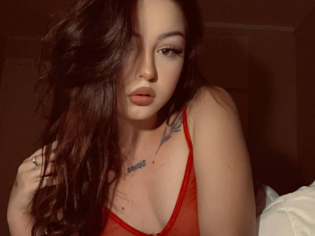 SinfulKitty with long, wavy brown hair, red lingerie, and a tattoo on her right shoulder, looking seductively at the camera with full lips and a slight smile.