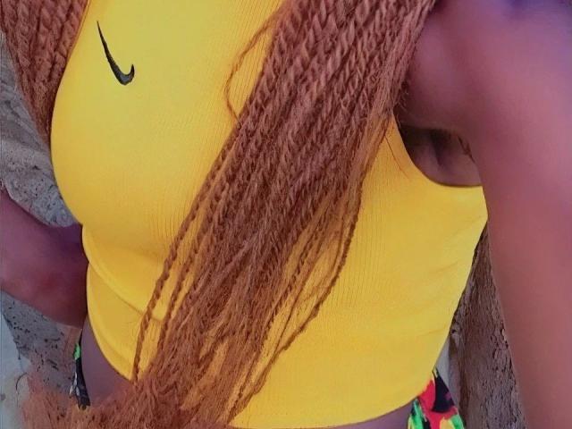 Sexyariaashley-sexy, usando uma blusa esportiva amarela da Nike que mostra seus seios, calças coloridas e tranças longas, finas e acastanhadas. Seu braço direito está parcialmente visível..