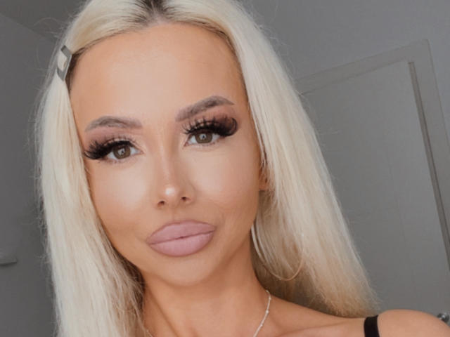 MoniqueBliss hat blondes Haar, volle Lippen mit pinkem Lippenstift und dicke, lange Wimpern. Ihre Brüste sind klein und ihr Hintern ist nicht sichtbar. Sie trägt einen schwarzen Träger..