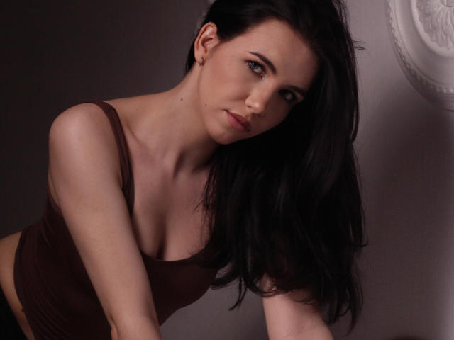 Tricia Barnes, met donkerbruin haar, draagt een bruin tanktop, leunt voorover, toont haar kleine borsten en heeft een lichte glimlach, met een serieuze, sexy en schattige uitdrukking..