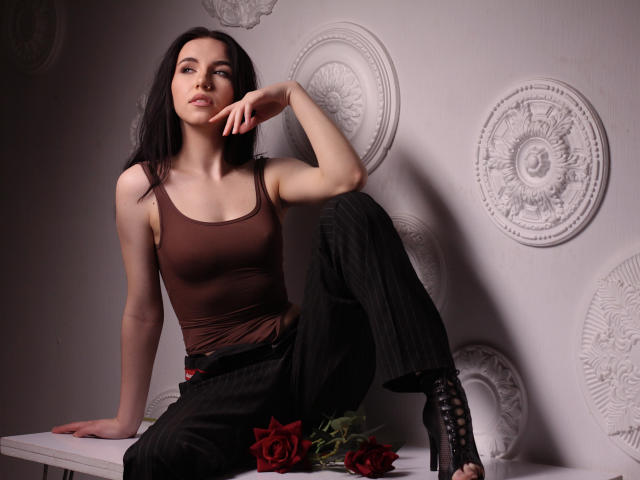 Tricia Barnes, avec ses cheveux foncés, son débardeur marron, son pantalon noir à rayures fines, ses bottes noires à lacets, assise avec une jambe levée, la main sur le menton, avec des roses rouges devant elle..