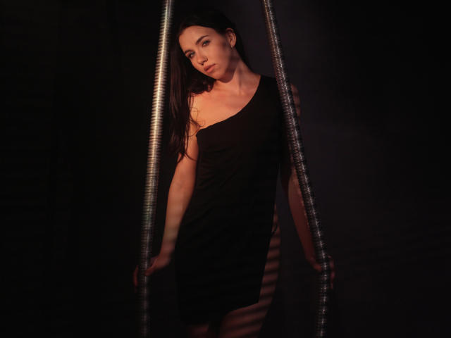 TriciaBarnes with long dark hair, wearing a black one-shoulder dress, standing in a dark, metallic, pipe-like structure, looking slightly to the side with soft, ambient lighting.
