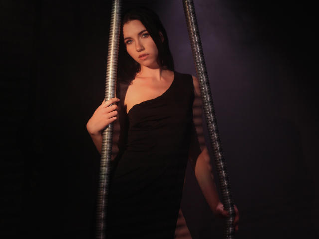 Tricia Barnes, avec ses cheveux bruns foncés, portant une robe noire à une épaule, se tient entre deux poteaux métalliques, en tenant l'un d'eux avec une expression sérieuse, dans une lumière tamisée, son postérieur étant légèrement visible..