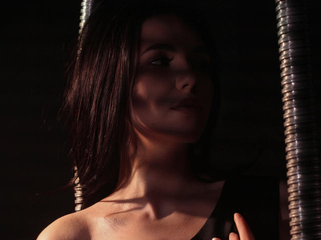 Tricia Barnes, com cabelo castanho escuro, sorriso suave e ombros à mostra, parcialmente obscurecida por postes metálicos, realçando suas características sutis, sensuais e atraentes..