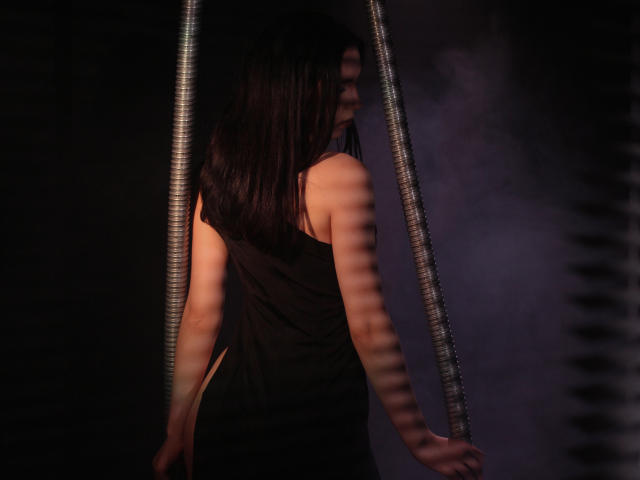 TriciaBarnes with dark, straight hair, wearing a black, backless dress, standing in shadow with striped light pattern on her back, looking over shoulder, dimly lit.