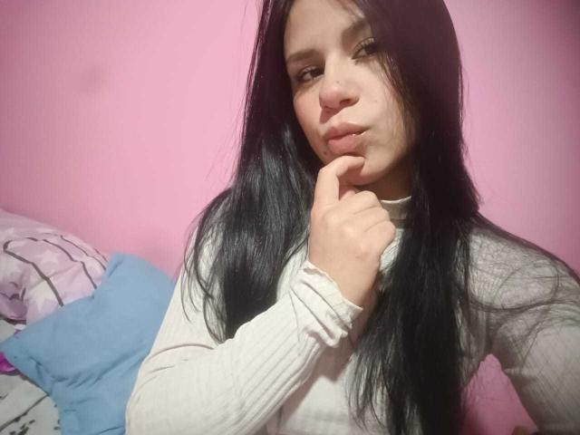 GinaRider with long black hair, pouty lips, and finger touching her chin, wearing a white ribbed top, pink background, and blue pillow in the lower left.