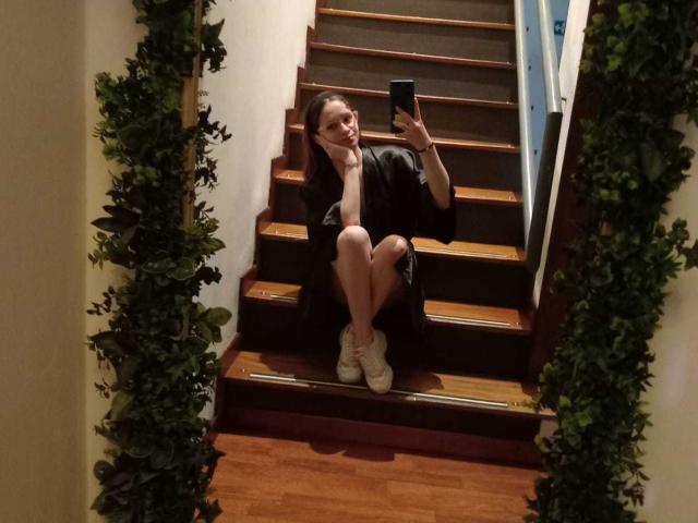 LesleyGray, zittend op houten trappen, gekleed in een zwarte mantel, witte sneakers en zonder ondergoed, maakt een selfie met een telefoon, waarbij haar blote achterwerk te zien is..