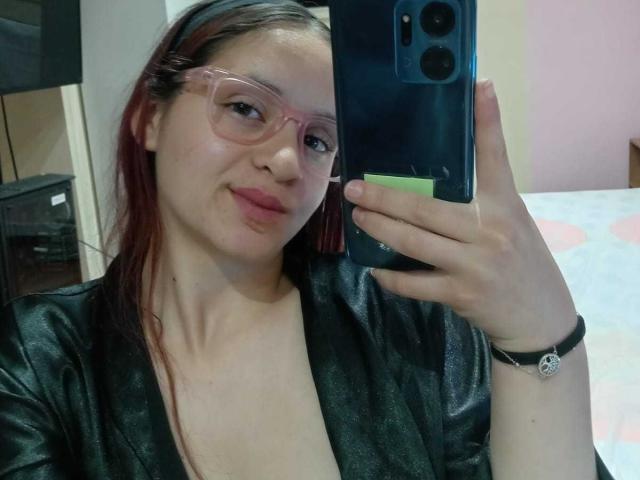 LesleyGray met roze bril, rood haar, een zwarte leren mantel, die een telefoon vasthoudt, lachend, met een schattig gezicht, kleine borsten, lichte huid, roze lippenstift, een pols met een zilveren armband..
