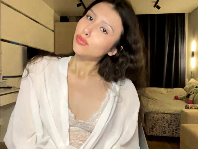 NecoleArciniega with wavy dark hair, pale skin, and red lipstick, wearing a white robe over a lace bra, pouting slightly, in a dimly lit room.