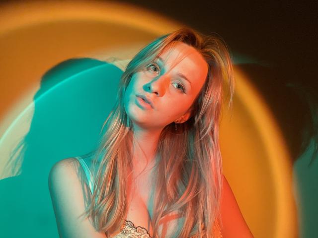 FlyFreedom with blonde hair, blue-green and orange light, slightly tilted head, subtle smile, small earrings, and visible cleavage, looking directly at camera.