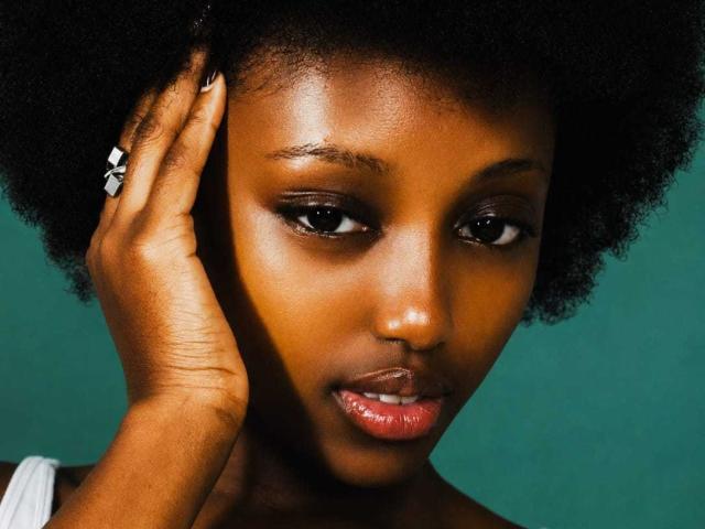 O rosto marrom escuro de KeishaJoy, com lábios brilhantes, cabelo afro e um anel branco no dedo esquerdo, tocando sua têmpora. Sua pele é lisa e ela tem uma expressão séria..