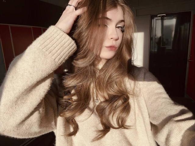 LauraleePenderel with long, wavy brown hair, wearing a beige sweater, sunlight in her face, hand in her hair, soft expression, subtle makeup, natural light, relaxed vibe.