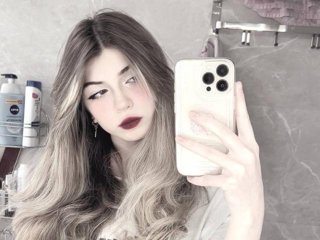 LauraleePenderel with long, wavy, blonde hair, holding a white iPhone, taking a selfie, wearing dark red lipstick, in a bathroom with toiletries and a towel.