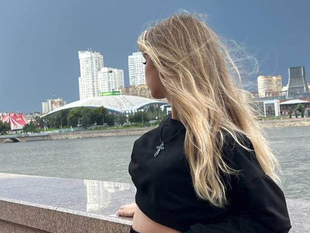 LauraleePenderel with long blonde hair, black top, and necklace, standing by a waterway, looking away, city skyline in background, wearing glasses, holding a black object.