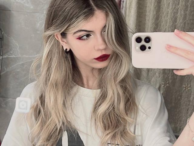 LauraleePenderel with long blonde hair, red lipstick, white shirt, black ear gauges, holding pink iPhone, taking selfie, looking at camera, neutral expression, grey tiled wall.
