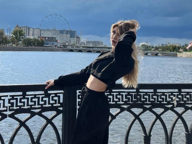 LauraleePenderel with long blonde hair, leaning on a black railing, wearing a black crop top and high-waisted black pants, standing by a river with a Ferris wheel in background.